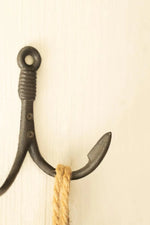 A rustic black cast iron anchor wall hook with a textured grip and a natural tan rope wrapped around its shank, set against a light cream-colored wooden background.