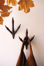 Two dark brown ceramic chicken feet wall hooks hanging on a white wall, with dried autumn leaves at the top and brown fabric bags hanging from the hooks below.