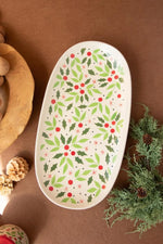An oval ceramic serving platter with a white background decorated with a green holly and red berry pattern, styled on a brown surface with holiday props.