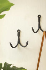 Two black metal anchor-shaped wall hooks mounted on a light cream wall, with green leaves at the corners and a wooden stick hanging on the right.