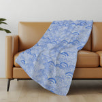Blue patterned blanket draped over a brown sofa in a room with a light-colored wall and wooden floor.