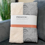 Two folded blankets, one cream and one gray with shell pattern, on a couch with 'Premium Washables' label.