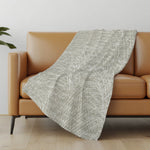 Gray textured blanket draped over a brown sofa in a room with a light gray wall and wooden floor.