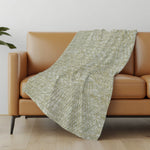 Green patterned blanket draped over a brown sofa in a room with a light-colored wall and wooden floor.