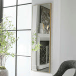 Tall floor mirror with gold frame leaning against a white wall next to a window.