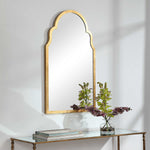 Gold-framed mirror on a white wall with a glass table below holding books and a vase with flowers.