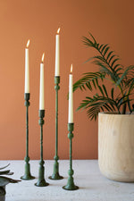 Green candlesticks with lit candles on a table against an orange wall with a plant in the background.