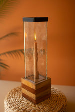 Candle in a glass holder with wooden base on a textured surface against an orange wall.
