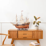 Wooden model ship on a wooden sideboard with a plant and books against a white wall.