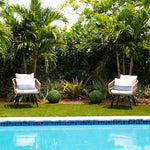 Two wicker chairs with white cushions by a pool surrounded by greenery