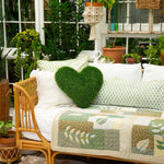 A cozy outdoor daybed setup with a green textured heart-shaped throw pillow, white and patterned pillows, and a green and beige patchwork quilted blanket, surrounded by lush plants and a rattan chair.