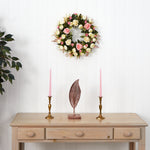 Decorative wreath with flowers on a white wall above a wooden table with candles and a leaf sculpture.