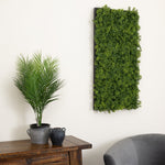A rectangular artificial green foliage wall hanging mounted on a white wall, shown in a styled interior with a potted palm on a wooden sideboard and part of a gray chair visible.