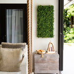 A rectangular green artificial hedge wall hanging mounted on a white wall in a bright porch or patio setting, with a wooden side table below holding a bowl and small decor items.