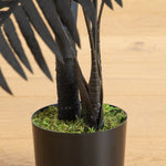 A potted artificial palm tree with black faux fronds and a textured black trunk, set in a black pot filled with green moss, placed on a light wood floor.