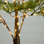 Warm white LED fairy string lights wrapped around a decorative tree trunk, with green foliage visible against a soft gray background.