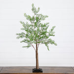 An artificial cypress tree with green foliage and a brown trunk, displayed on a black square base over a wooden table against a white shiplap wall.