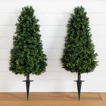 Two artificial topiary trees on stands against a white wooden panel background