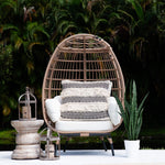 A woven rattan egg-shaped outdoor chair with white cushions and a black-and-white patterned throw pillow, styled on a patio with a potted snake plant, lanterns, and a lush tropical background.