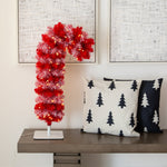 A red artificial Christmas tree with white tinsel-like accents and warm white string lights, shaped like a candy cane, displayed on a wooden console table next to two decorative throw pillows against a cream wall with framed abstract art.