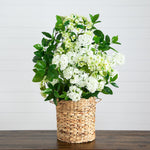 A faux floral arrangement of white flowers and green leaves in a woven natural fiber basket, displayed on a dark wooden surface against a white paneled wall.