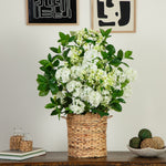 A potted artificial flowering plant with dense clusters of white blossoms and glossy green leaves, arranged in a woven natural-fiber basket on a wooden table against a light wall.