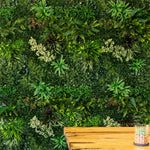 A lush artificial green wall mural featuring various faux ferns, moss, and small plants, with a wooden tabletop and a small glass planter visible in the bottom right corner.