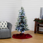 A flocked artificial Christmas tree with white lights and pinecones stands on a red velvet tree skirt in a living room corner with a gray armchair and wooden side table.