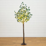 A tall artificial eucalyptus tree with glowing warm white LED fairy lights woven through the green leaves, standing on a wooden floor against a white shiplap wall.