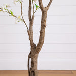 A decorative faux tree branch arrangement with textured brown bark, small green leaves, and white blossoms, shown against a white paneled wall and light wood floor.