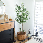 A tall artificial green plant in a woven wicker basket planter, styled in a bright bedroom next to a wooden and black dresser.