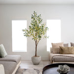 A tall potted artificial olive tree with green and yellow leaves stands in a bright, modern living room between two large windows, flanked by gray sofas with neutral-toned cushions.