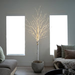 A decorative artificial tree with a white faux-birch trunk and delicate warm white string lights, displayed in a textured gray pot in a modern living room setting.