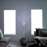 A decorative artificial tree with a white faux-birch trunk and many thin branches lit with multicolored LED fairy lights, placed in a gray woven pot in a modern living room setting.