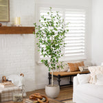 A tall artificial green tree in a textured white pot, styled in a bright, modern living room next to a white brick fireplace and white shutters.