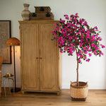 A potted artificial bougainvillea tree with bright pink flowers and green leaves, styled in a wicker basket next to a tall wooden cabinet in a bright room.