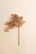 A single dried decorative branch with clustered small leaves in warm beige, tan, and muted rust tones, attached to a long brown stem against a plain cream background.