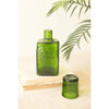 A green glass bottle with a matching green cap placed beside it on a light surface, with a book and palm leaves in the background.