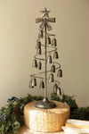 A rustic metal spiral Christmas tree ornament stand with hanging bells and a star topper, displayed on a woven round base with holiday greenery and books at the bottom.