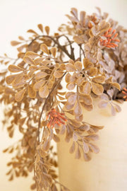 Artificial olive branch garland with muted brown and beige leaves and small orange-red buds, draped over a cream-colored vase against a warm off-white background.