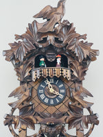 Wooden cuckoo clock with intricate carvings and two figures inside on a white background
