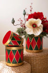 Two decorative cylindrical vases with a red and green harlequin diamond pattern and gold trim, styled with flowers on woven wicker stools against a neutral background.