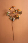 A dried floral arrangement featuring orange and yellow sunflower-like blooms with brown and gray leaves on long stems, set against a solid warm terracotta background.