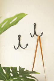 Two black wall hooks on a white wall with green leaves in the foreground