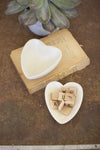 Heart-shaped ceramic dish with wooden letters on a textured surface with a plant in the background