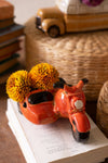 Orange scooter-shaped vase with yellow flowers on an open book, with a blurred background
