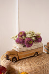 Decorative model of a bus with flowers on top, placed on a woven surface.
