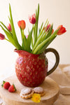 Red pitcher with tulips on a wooden surface with cookies and strawberries.