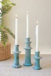 Three turquoise candle holders with white candles on a light surface, with a plant in the background.