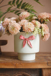 White vase with pink bow and floral arrangement on a wooden surface